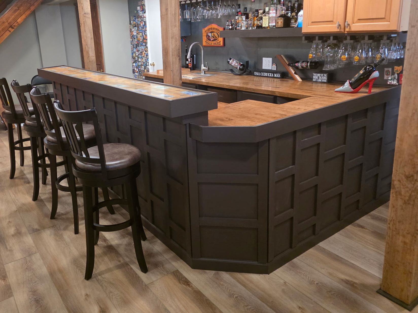 An L-shaped basement bar with a dark paneled front, wood countertop and four bar stools.