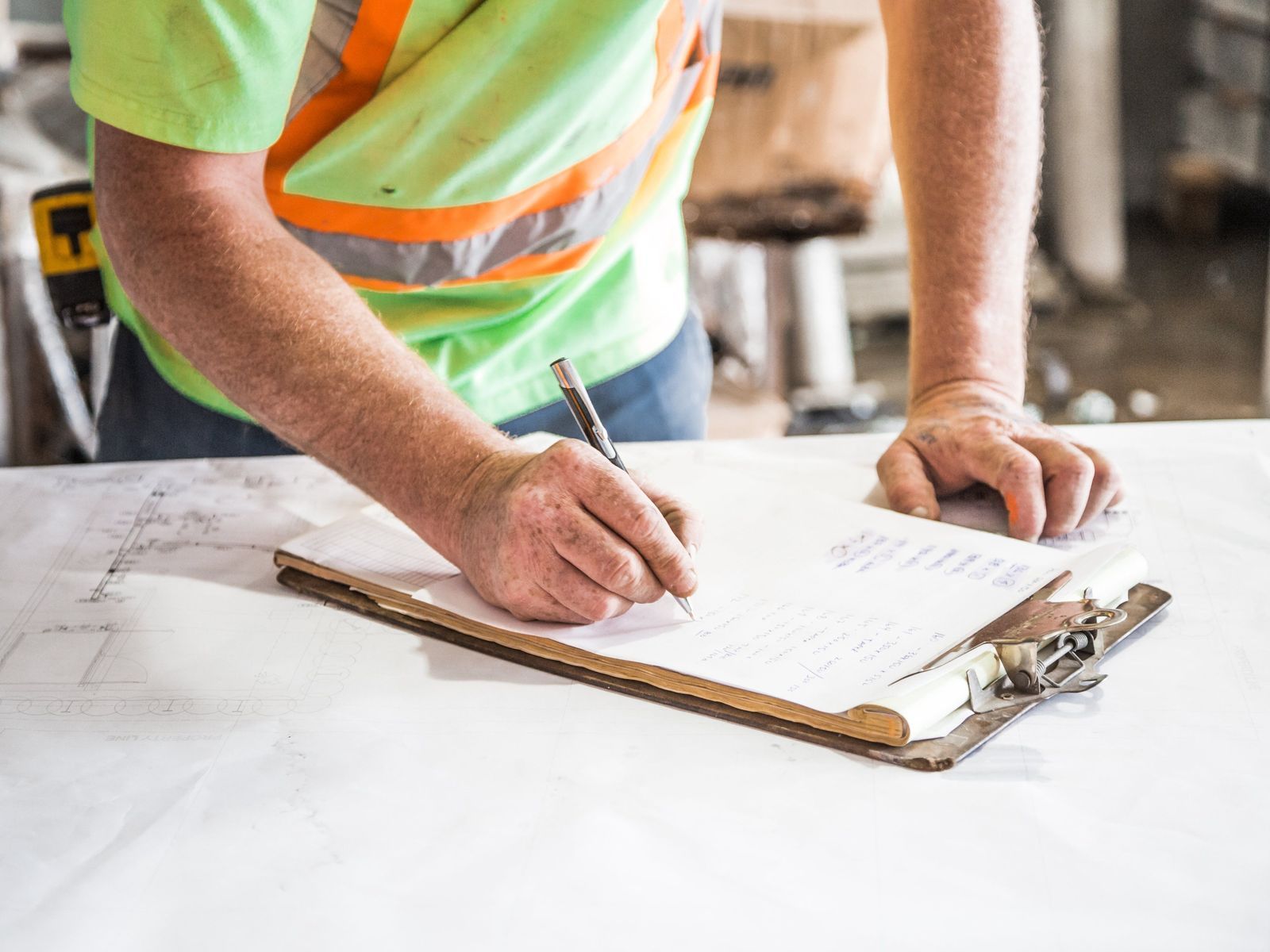 A worker in a high-visibility vest writing notes on a clipboard over a set of construction plans.