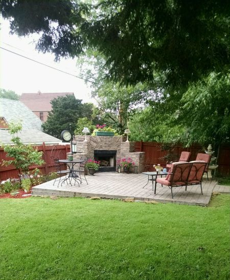 Backyard paver patio with a stone outdoor fireplace, seating and a lawn in front.